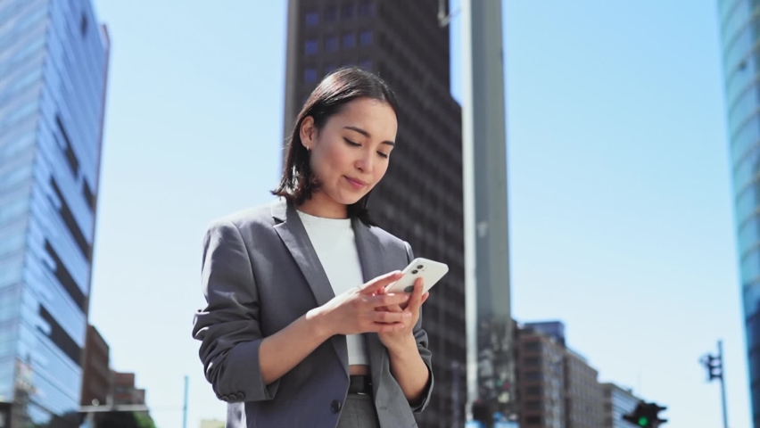 Successful elegant young Asian businesswoman wearing suit standing in big city talking on cell phone. Smiling woman making business call on cellular on sky background urban buildings, slow motion. Royalty-Free Stock Footage #1094083565