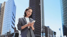 Successful elegant young Asian businesswoman wearing suit standing in big city talking on cell phone. Smiling woman making business call on cellular on sky background urban buildings, slow motion. - Powered by Shutterstock - Get 15% off with code: PIKWIZARD15