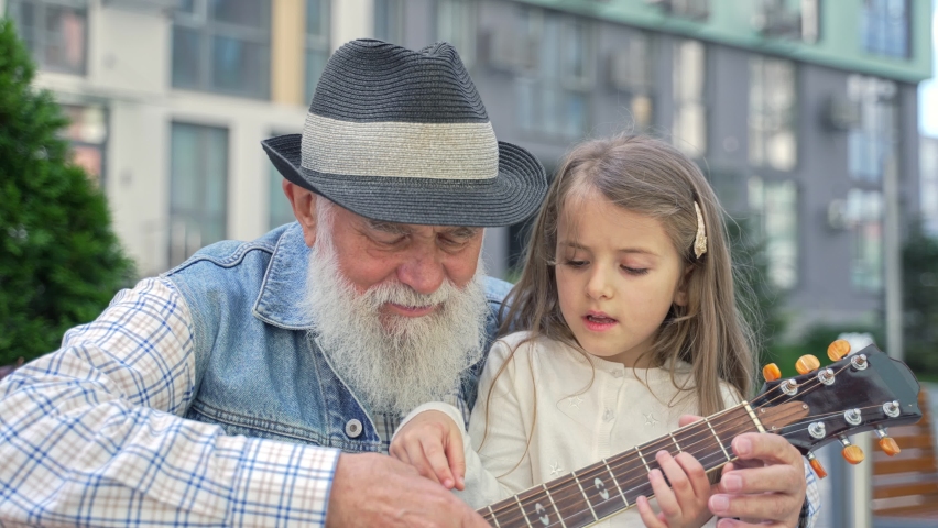 Close up caring senior caucasian grandpa teaching his happy lovely little granddaughter to play guitar sitting on a bench outdoor on the street. High quality 4K footage 