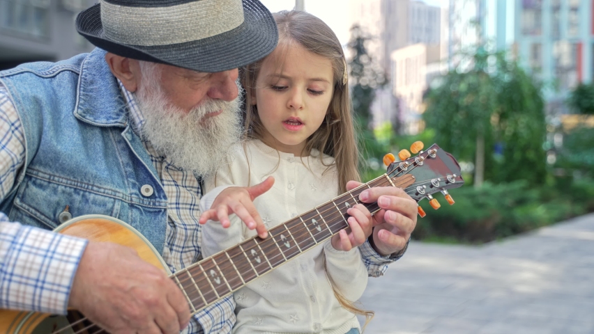 Zoom out caring senior caucasian grandpa teaching his lovely enthusiastic little granddaughter to play guitar sitting on a bench outdoor on the street. High quality 4K footage 