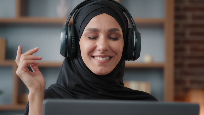 Muslim Islamic woman in black hijab wearing headphones headset listening musing song audio sound indoors with laptop computer radio app in online musical website moving dancing with hands front view Royalty-Free Stock Footage #1094091903