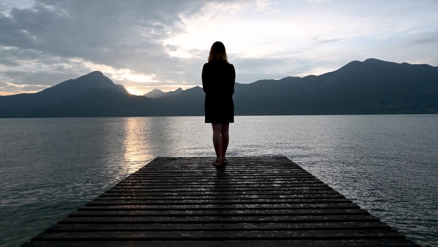 Girl standing at the end of the pier admiring the beautiful sunset, lake and the mountains. Tripod shot