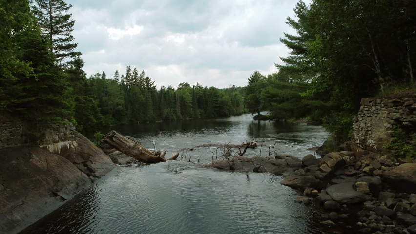 Aerial shot of the flowing Oxtongue River as the water passes through a rocky narrowing cascading over the edge of a waterfall pushing through trees and debris, Algonquin Provincial Park, Canada
