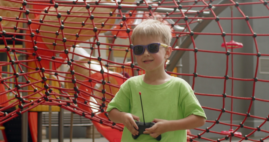 Swagging preschooler boy wearing bright sunglasses shows remote controller from toy car. Happy child enjoys walking on modern playground with toys