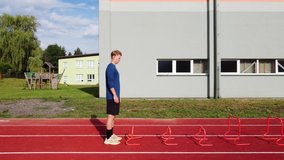 Blond athlete in a blue T-shirt develops his dynamic strength by jumping over hurdles on an athletic oval. 4k video