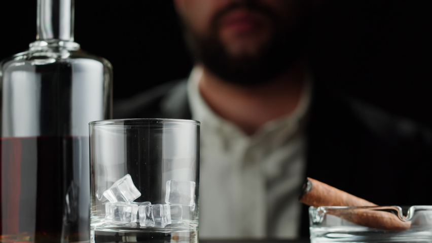 Drinking brandy or whiskey close-up. Rich man drinks luxury cognac in ice on black background. Alcohol amber rum, liqour beverage in glass. Smoking cigar. 