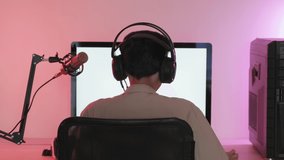 Back view of streamer in headset playing on video game on computer at desk with microphone in room lit with neon light
