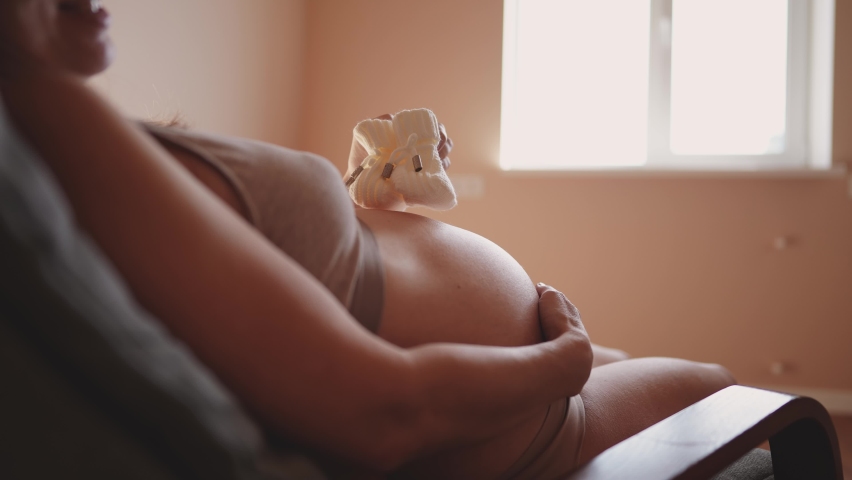 Pregnant woman with belly. woman with big bare a belly holding booties baby shoes indoor close-up. health pregnancy motherhood procreation concept. girl preparing for motherhood | Shutterstock HD Video #1094124037