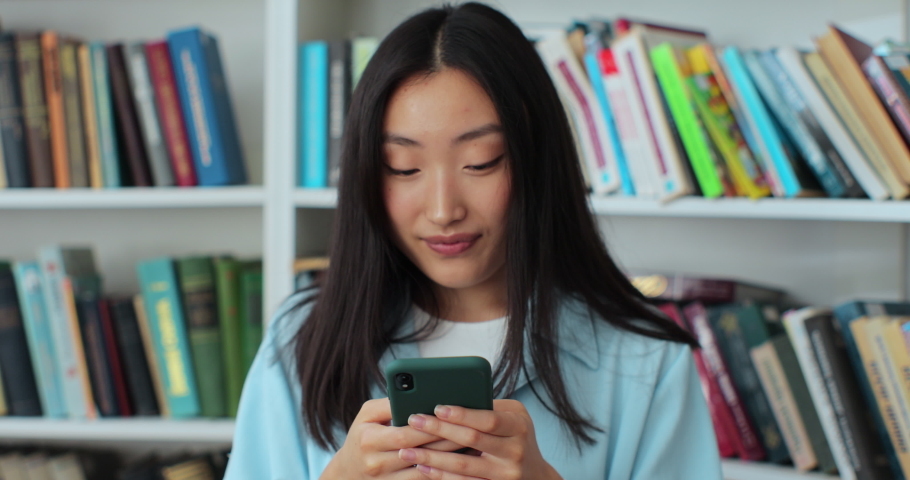 Attractive asian woman using smartphone, texting message while standing in the library.