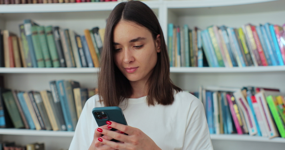 Student girl standing in library and using smartphone, texting message, check on-line studies timetable using mobile application.