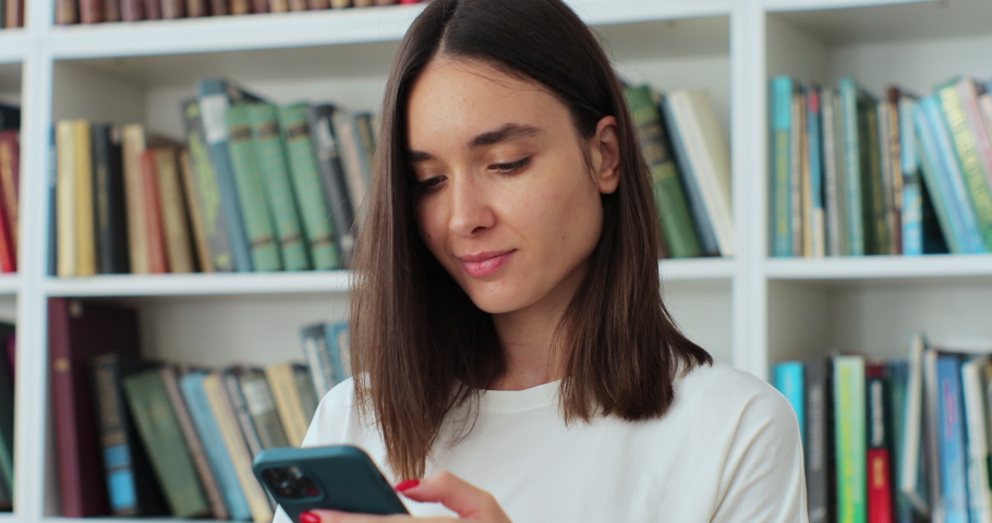 Close up student girl standing in library and using smartphone, texting message, check on-line studies timetable using mobile application.