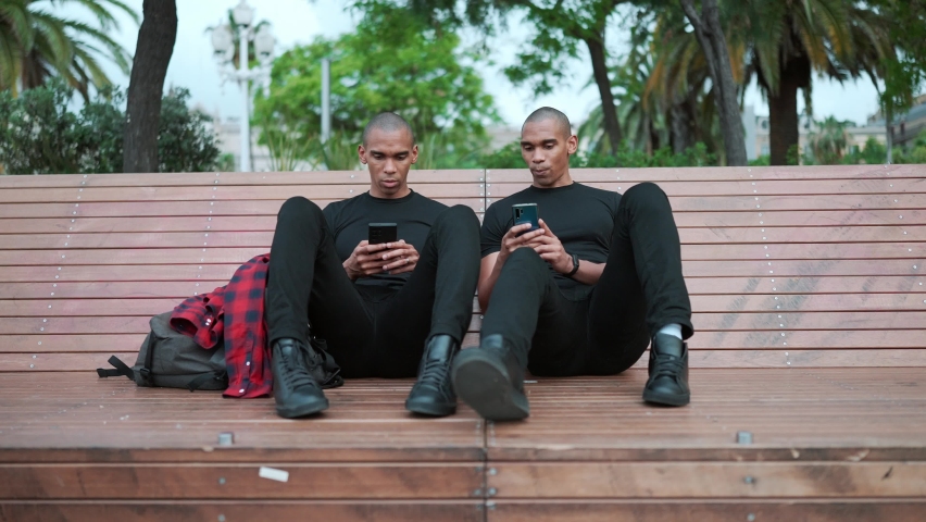 Two confident bald African brothers talking and typing on phone while sitting on the bench outdoors