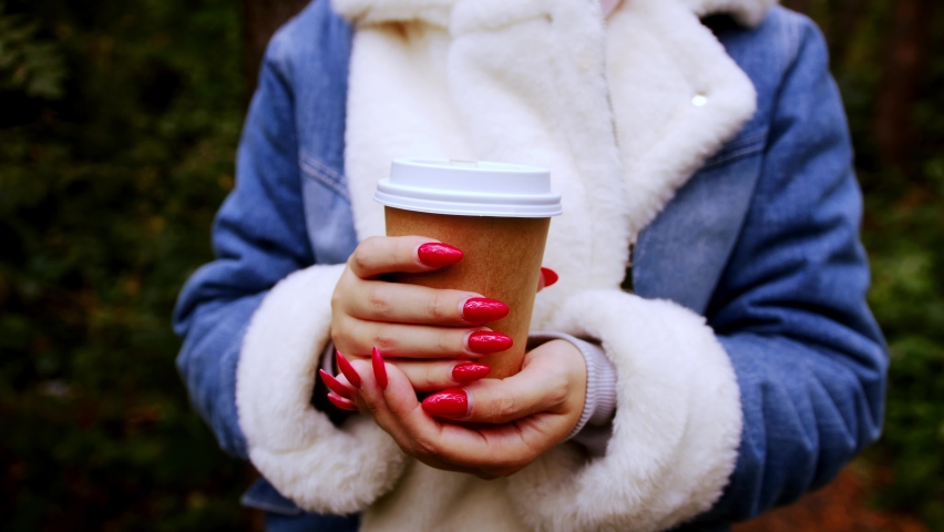 Paper cup with coffee in hands of unrecognizable woman, standing in forest. Body part of female in warm clothes with bright manicure holding hot drink.