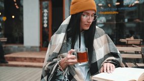 Smiling young brunette woman wearing hat and eyeglasses reading book and drinking tea in cafe outdoors - Powered by Shutterstock - Get 15% off with code: PIKWIZARD15