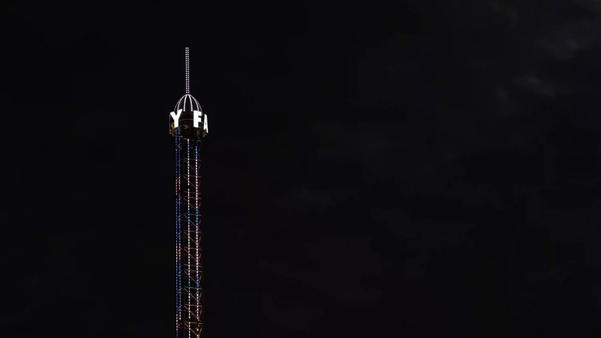 Close-up of rotating letters of a sky fall or free fall or high gravity or drop tower at night. Carnival, amusement or thrill or theme park entertainment. Colourful neon lights against the black sky.