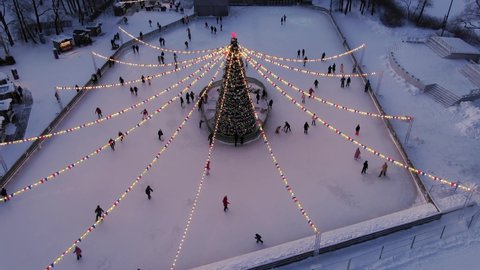 Festive city ice rink with people skating around Christmas tree with illuminating garlands and decorations in winter evening aerial panorama Stock Video