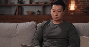 Asian man using laptop and working remotely from home, sitting on couch. Focused man freelancer browsing the internet and chatting online on portable computer during distant job