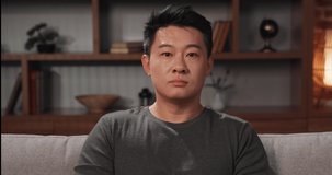 Portrait of Asian man looking at the camera at home. Focused man in casual cloth sitting on couch, participating in a video conference