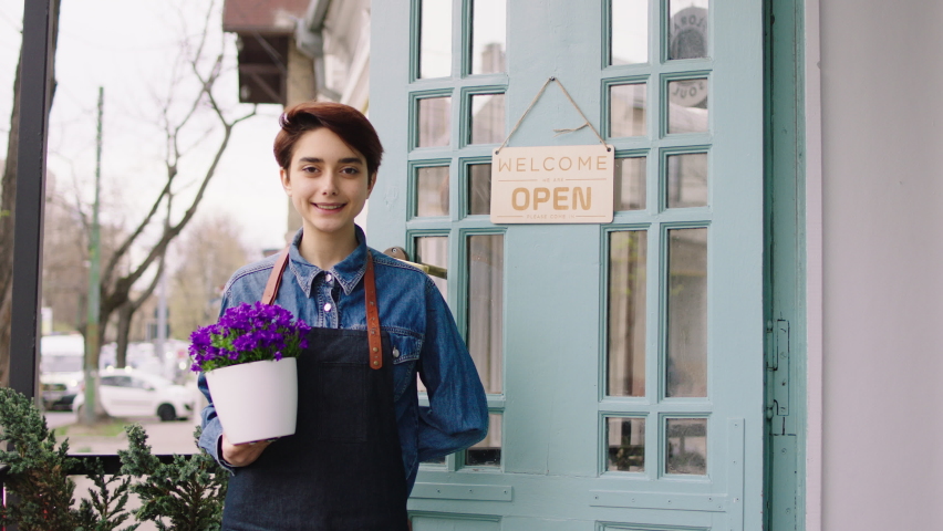In front of the camera posing with a flowers pot in hands a pretty florist lady she smiling large standing at the entry to the shop