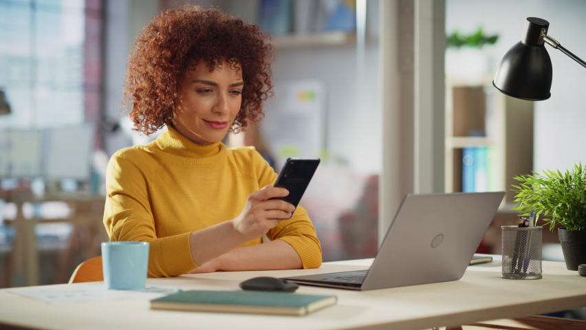 Beautiful Middle Eastern Manager Sitting at a Desk in Creative Office, Using Smartphone. Young Stylish Female Sending Message, Browsing Internet. Colleagues Working in the Background.