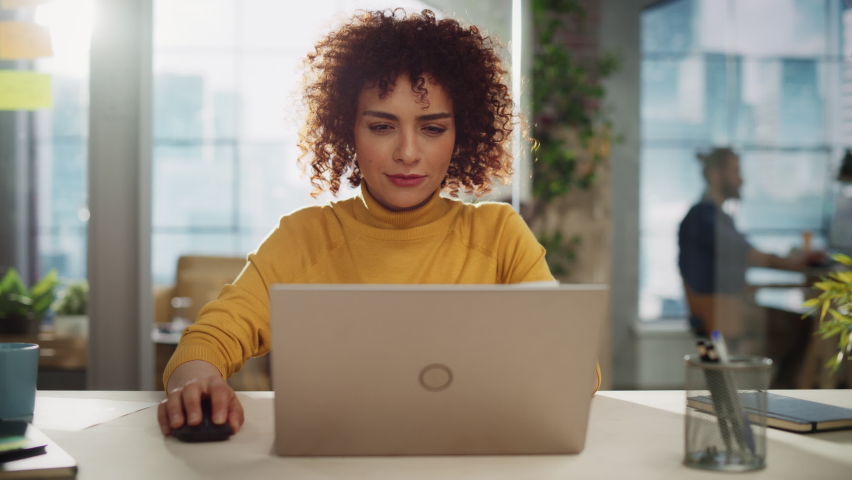 Portrait of a Beautiful Middle Eastern Manager Sitting at a Desk in Creative Office. Young Stylish Female with Curly Hair Looking at Camera with Big Smile. Colleagues Working in the Background. Royalty-Free Stock Footage #1094152395