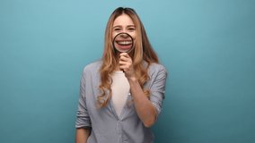 Smile, zoom, tooth care. Portrait of young woman standing and holding magnifying glass on her teeth, looking at camera, wearing striped shirt. Indoor studio shot isolated on blue background. - Powered by Shutterstock - Get 15% off with code: PIKWIZARD15