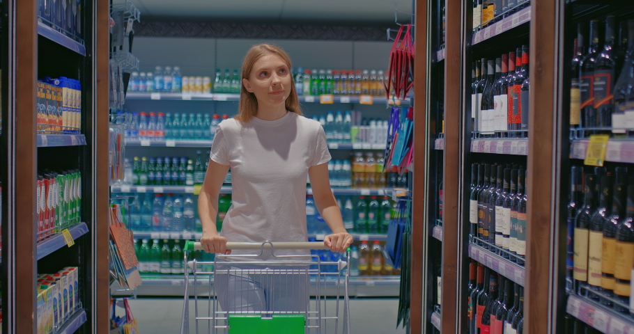 Beautiful girl in the department of alcoholic beverages chooses a bottle with a drink. Racks of alcoholic beverages stand in a row in the supermarket. 4k, ProRes