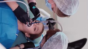 Vertical Video, Two Woman Doctors in Medical Cap in a Dental Clinic, Serve the Patient Little Boy with Damaged Baby Teeth. Work of a Dentist with Children. Healthcare and Medicine Concept.