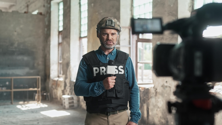 Male journalist correspondent wearing bulletproof vest and helmet reporting live in damaged building. Military concept. Behind the scenes of war. Man newsman speaking in microphone at camera. War zone Royalty-Free Stock Footage #1094171697