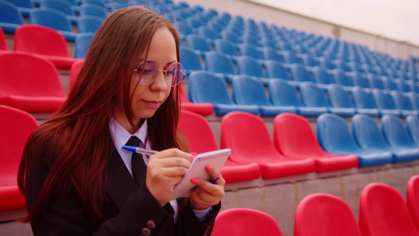 Young woman with notepad, pen sitting on stadium bleachers alone. Female journalist writing down notes during sports training at street stadium.