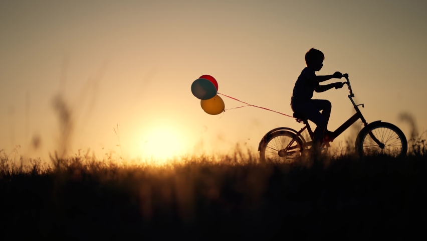 Dream child. Silhouette of kid on bike in park. Boy rides in park on green grass. Child games in nature.Traveling with balloons on bike.Active child freedom in summer.Boy learn to ride bike in nature. Royalty-Free Stock Footage #1094180311