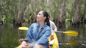 4K Young Asian couple traveler kayaking in mangrove forest on summer travel vacation. Man and woman tourist using mobile phone taking selfie or video call while paddling on canoe boat in forest lake