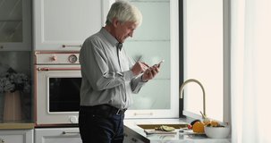 Focused mature grey haired man cooking salad in kitchen, consulting Internet, reading recipe, chatting, getting happy, smiling, using mobile phone at table with fresh vegetables ingredients