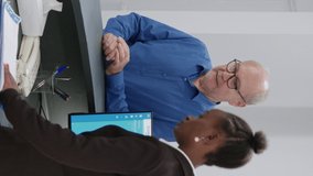 Vertical video: Old man analyzing checkup form papers at hospital reception counter, receiving help and support from receptionist. Patient signing medical report before healthcare appointment at desk.