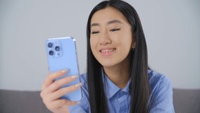 Happy Asian girl talking on video call. Cheerful Vietnamese female person calling her friends on frontal camera in modern blue smartphone with triple camera. BIPOC person lifestyle video clip 