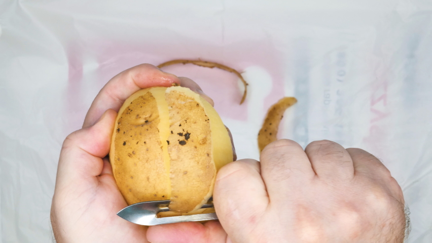 Peeling potatoes. Man's hands peeling potatoes. A man peels raw fresh potatoes with a manual potato peeler. Peeled potatoes and peel. Cooking food in the kitchen. Vegetarian food. Close-up. Top view