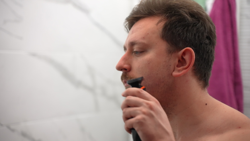 Handsome man in profile shaving cheeks, moustache and beard with electric trimmer in the bathroom against colourful towels background, young male doing daily skin care treatment at home. Person