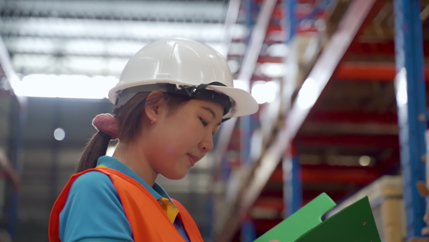 Asian female employees, Wear white helmet, wear orange reflective vest, for safety while working, He was standing to check packaged goods in order deliver the next step, he holds a green checkbook