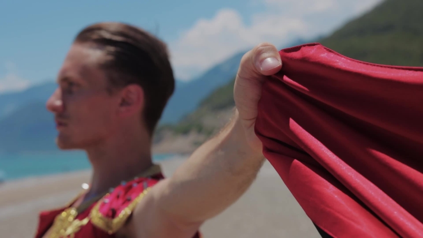 An attractive man in a spanish suit stands by the sea and holds a red robe