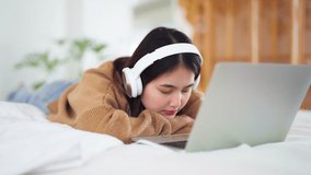 Happy beautiful young asian woman working on computer laptop and lying down on bed in house. She making video conference webinar or wearing headphone listening music