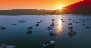 Sun sending last rays from behind the mountains. Multiple yachts and boats standing on anchor on the lake. Orange sky at backdrop.