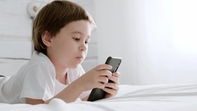 4K. Little baby boy lays on his bed and plays with a smartphone. Close-up portrait. Holds in hands black cell phone. Video footage. Concept of online education. White room and clothes. Telephone user.