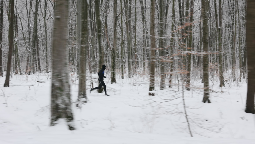 Active young guy dressed in black sport clothes spending morning time for jogging outdoors. Caucasian sporty man enjoying leisure activity among forest with snow lying around.