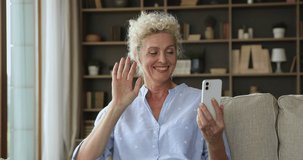 Happy pretty senior mobile phone user woman waving hand hello at screen, frontal camera, smiling, saying hi, speaking on video conference call, talking to family online, laughing, sitting on home sofa