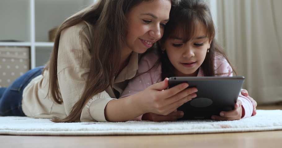 Happy young mom and daughter kid using online learning app on tablet, lying on heating carpeted floor at home, holding gadget, laughing, smiling, shopping on Internet, playing video game Royalty-Free Stock Footage #1094244507