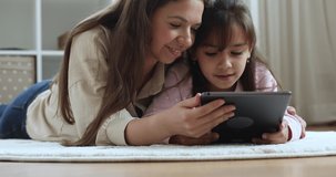 Happy young mom and daughter kid using online learning app on tablet, lying on heating carpeted floor at home, holding gadget, laughing, smiling, shopping on Internet, playing video game