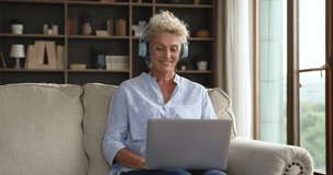 Happy friendly senior woman in big bright wireless headphones talking on video call on laptop, waving hand hello at webcam, speaking, smiling, listening, nodding, sitting on couch at home