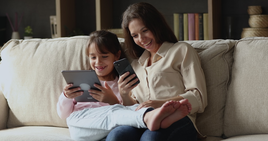 Happy mom and gen Z daughter kid using smartphone and tablet, relaxing on couch together, talking, laughing, sharing media content. Mother teaching girl to use app on internet Royalty-Free Stock Footage #1094244571