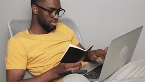 African american man in glasses looking in a notebook student and working in a laptop checking his knowledge at home online quarantine online learning