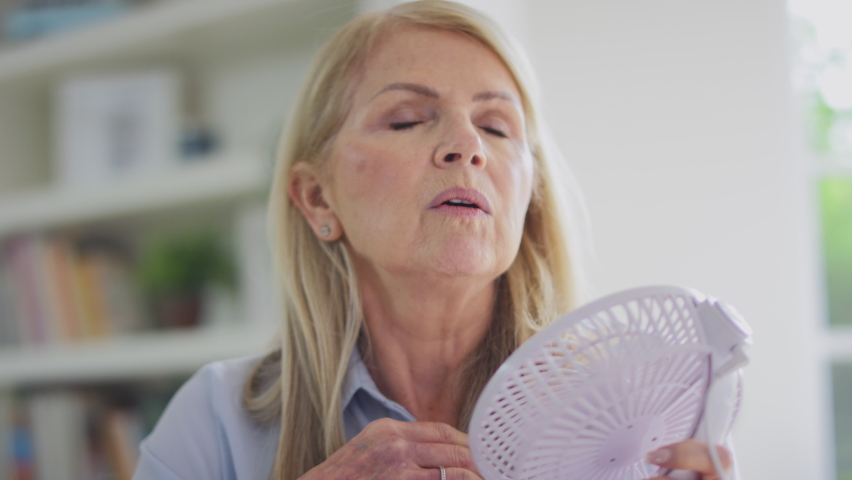 Menopausal mature woman at home having hot flush and cooling herself with electric fan shot in slow motion Royalty-Free Stock Footage #1094252255